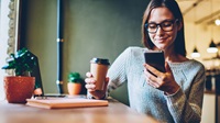 Kvinde sidder i sit køkken, nyder en kop kaffe og tjekker udviklingen på sine fonde i mobilbanken på sin telefon