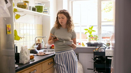 ung kvinde i køkken tjekker sin månedsinvestering på mobiltelefonen